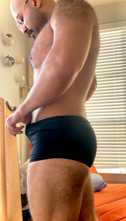 Person wearing black underwear in a room with a bed and window blinds.